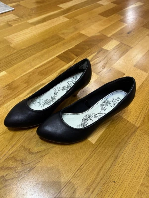 Svarta ballerinaskor med klack - Snygga svarta ballerinaskor från Vion Shoes i storlek 39. Skorna har en låg klack och en spetsig tå, tillverkade i skinnimitation. Insidan har ett blommigt mönster i svart och vitt som ger en extra detalj. Perfekta för dig som gillar stilrena och klassiska skor.