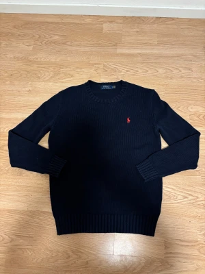 Mörkblå stickad tröja Polo Ralph Lauren - Snygg mörkblå stickad tröja från Polo Ralph Lauren med klassisk röd logobrodering på bröstet. Rund halsringning och ribbade muddar vid ärmslut och nederkant. Perfekt för dig som gillar stilrena och tidlösa plagg.