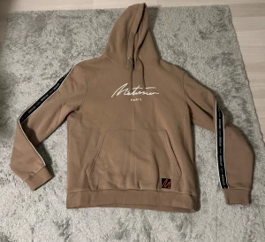 Beige hoodie från Métissier Paris XL - Snygg beige hoodie från Métissier Paris i storlek XL. Tröjan har huva med snörning, vit logotyp på bröstet och svarta band med text längs ärmarna. Praktisk magficka och en cool detalj med orange broderad M på nederkanten. Perfekt för chill dagar.