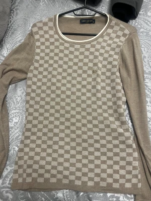 Beige rutig tröja  Louis Vuitton - Snygg beige långärmad tröja från Louis Vuitton med rutigt mönster på framsidan och diskret LV-logga. Tröjan har rund hals med vit kant och är tillverkad i mjukt stickat material som känns lyxigt och skönt mot huden.