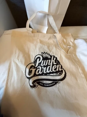 Beige shoppingväska Punk Garden - Snygg beige shoppingväska från Punk Garden med svart tryck och coolt grafiskt motiv på framsidan. Väskan har två handtag och är tillverkad i slitstark bomull, perfekt för att bära med sig allt man behöver under dagen.
