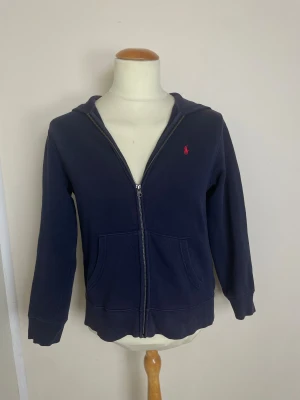 Marinblå hoodie från Polo Ralph Lauren - Snygg marinblå hoodie från Polo Ralph Lauren med röd broderad logga på bröstet. Tröjan har huva, dragkedja framtill och två praktiska fickor. Tillverkad i mjuk bomull, perfekt för chill dagar. Klassisk och stilren design som funkar året runt.