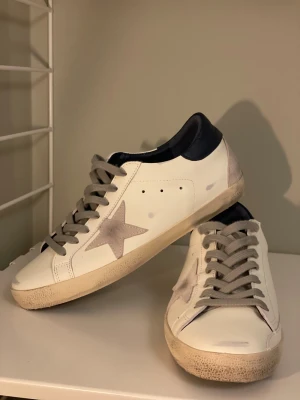 Golden Goose vita sneakers med stjärna - Snygga vita sneakers från Golden Goose med grå mockadetaljer och en ikonisk stjärna på sidan. Skorna har grå snörning, mörkblå häl och platt sula. Perfekta för dig som gillar en cool och avslappnad stil med unika detaljer.ps boxen medföljer 