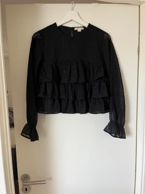 Svart blus med volanger - Säljer en svart blus från gina med glittriga prickar och flera lager volanger framtill. Blusen har långärmade transparenta ärmar med volang vid ärmslut och en rund halsringning. Perfekt för dig som gillar detaljer och en lite extra look.