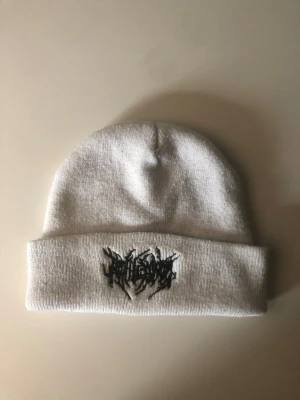 Vit stickad mössa med svart brodyr - Vit mössa i stickad material med uppvikt kant och svart broderad logga framtill i ett edgy, grafiskt typsnitt. Perfekt för dig som gillar streetwear och vill ha en clean men ändå unik look. Passar till både chill och lite kallare dagar.