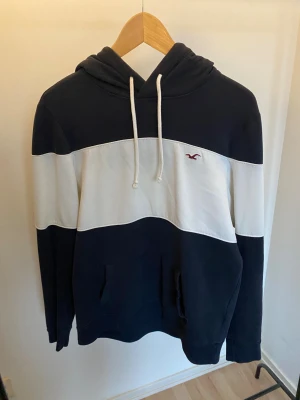 Blåvit hoodie från Hollister -storlek small - Snygg hoodie från Hollister i storlek S med breda marinblå och vita blockränder. Tröjan har huva med snörning, känguruficka och lång ärm. Materialet är mjuk bomull och polyester, perfekt för chill dagar. Diskret Hollister-logga broderad på bröstet.