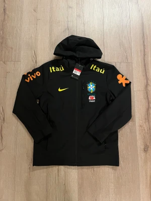 Svart Nike Brasil vindjacka med huva - Svart Nike vindjacka med huva, Brasil CBF-logga och sponsormärken i gult och orange. Jackan har hel dragkedja, lång ärm och är tillverkad i lätt polyester med Dri-FIT och Breathe-teknologi. Perfekt för fotboll och streetwear.