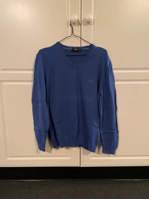 Blå v-ringad Hugo Boss tröja - Säljer denna kornblå stickade Hugo Boss tröja i storlek XL, skulle dock säga att den även passar L. Den är v-ringad och gjord av högkvalitativt italienskt ull. Den är i hyfsat skick. Tror inte att denna modell säljs längre men liknande modeller ligger på runt 1400kr. Hör av er vid eventuella funderingar!