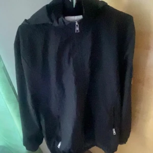 Svart vindjacka från Moncler - Svart vindjacka från Moncler med huva och dragkedja framtill. Jackan har två fickor med dragkedja och Moncler-logga på ärmen. Materialet är lätt och syntetiskt, perfekt för blåsiga dagar. Stilen är clean och passar dig som gillar streetwear.