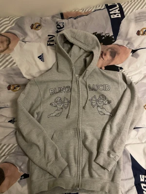 Grå hoodie från Blinglucid med änglar - Säljer en grå hoodie från Blinglucid i storlek S. Tröjan har dragkedja, huva med snörning och två fickor fram. På bröstet finns coola strassdetaljer med texten BLINGLUCID och två änglar. Materialet är mjuk bomull och polyester.