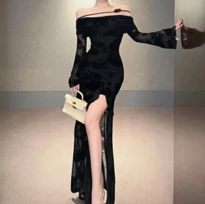 Svart långklänning med slits och offshoulder blomma  - Supersnygg svart långklänning med offshoulder och transparenta långärmar. Klänningen har en blommig mönsterstruktur, en hög slits framtill och en dekorativ svart blomma vid axeln. Materialet är lätt och har en lyxig känsla, perfekt för att sticka ut.