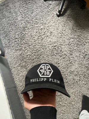 Svart Philipp Plein keps med brodyr - Snygg svart keps från Philipp Plein med vit broderad logga och text framtill. Klassisk böjd skärm och justerbar passform. Tillverkad i bomull för skön känsla och stilren look. Perfekt för dig som vill sticka ut med ett exklusivt streetwear-plagg.