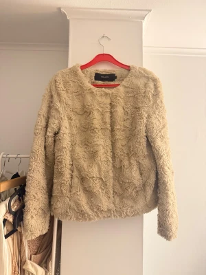Beige pälsjacka - Pälsjacka i beige från Vero Moda. Jackan har rund halsringning, långa ärmar och stängs med tryckknappar framtill. 
