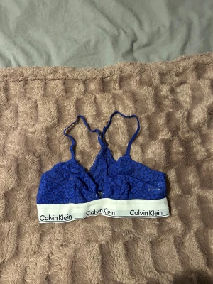 Blå spets linnetopp/bh Calvin Klein - Säljer en snygg blå linnetopp/bh från Calvin Klein med spetsdetaljer och elastiskt vitt band med logga. Toppen har smala axelband och är genombruten med blommigt spetsmönster. Perfekt för dig som gillar sportig och trendig stil.