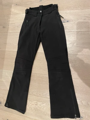 Svarta skidbyxor från McKinley, strl 158 - Svarta skidbyxor från McKinley med justerbar midja, dragkedjor vid fickor och benslut samt förstärkta knän. Byxorna är bootcut och  lite mer åt slim-fit hållet. Fleece-fodrade.  