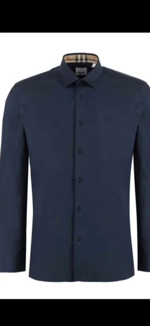 Mörkblå Burberry skjorta med rutiga detaljer - Snygg mörkblå skjorta från Burberry med klassiska rutiga detaljer på insidan av kragen och ärmsluten. Skjortan har lång ärm, knappar framtill och en stilren passform. Perfekt för dig som vill ha en clean look med en touch av lyx.