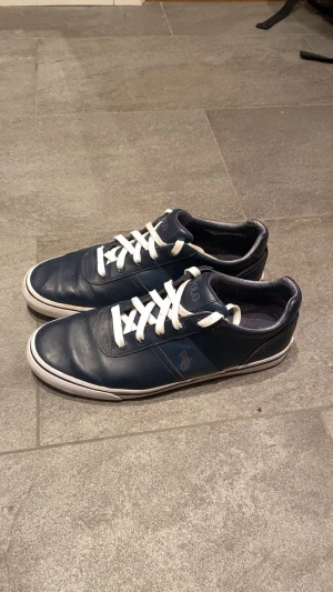 Mörkblå sneakers Polo Ralph Lauren - Snygga mörkblå sneakers från Polo Ralph Lauren med vit sula och vita snören. Skorna har klassisk låg profil, rund tå och är tillverkade i skinn. Polo-logga på sidan och märkesdetaljer på plös och innersula. Perfekta för en clean och stilren look.