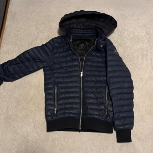 Mörkblå Moose Knuckles dunjacka - Säljer en mörkblå dunjacka från Moose Knuckles med huva och quiltad design. Jackan har dragkedja framtill, två sidofickor med dragkedja och ribbade muddar. Materialet är dun och återvunnen polyester, perfekt för kalla dagar. Stilsäker logga på ärmen.