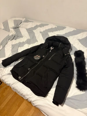 Svart Moose Knuckles Vinterjacka - Säljer en svart dunjacka från Moose Knuckles med avtagbar pälskrage, stora logodetaljer på ärmen och innerfoder med broderad hockeygrafik. Jackan har dragkedja, tryckknappar och flera fickor framtill. Perfekt för kalla vinterdagar.
