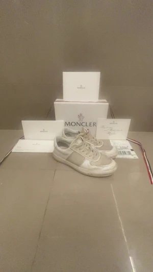 Vita sneakers från Moncler - Snygga vita sneakers från Moncler med klassisk låg profil och rund tå. Skorna har snörning och är tillverkade i skinn med detaljer i vitt och en diskret Moncler-logga på hälen. Perfekt för dig som gillar stilrena och exklusiva sneakers.