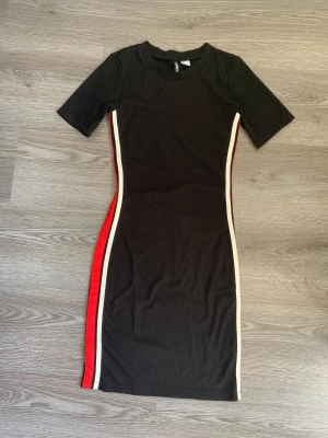 Svart tight klänning från H&M - Svart kortärmad klänning från H&M i storlek 36. Modellen är tight och har röda och vita ränder längs sidorna som ger en sportig vibe. Klänningen är i mjuk bomull och har rund hals. Perfekt för dig som gillar en enkel men snygg look.