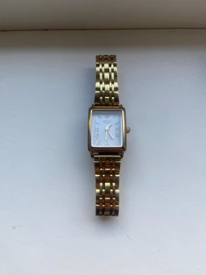 Guldfärgad fyrkantig klocka från Regal - Snygg guldfärgad klocka från Regal med fyrkantigt vitt ur och romerska siffror. Länken är i metall med klassisk design och har ett smidigt spänne. Perfekt accessoar för dig som gillar stilrena detaljer och tidlös look.