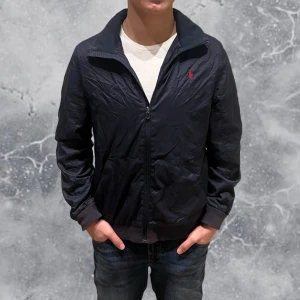 Ralph Lauren Windbreaker jacka navy blå - •Ralph Lauren Windbreaker Jacka mörkblå. Skick: 7-8/10, liten defekt vid ena fickan, inte märkvärdigt (se sista bild). Annars i helhet väldigt fint skick! Storlek: XL 18-20 ungdom storlek = XS~. •Mått, axel-neråt: 58 cm, bredd: 48 cm. Modell: 173 cm, 63 kg 