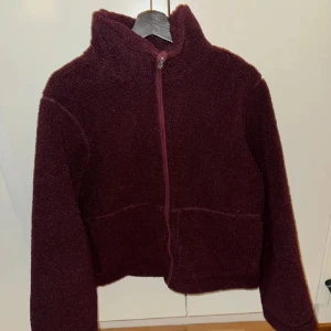 Vinröd teddyjacka med dragkedja - Mysig vinröd teddyjacka med hög krage och hel dragkedja framtill. Jackan har en fluffig textur och långärmad passform, perfekt för kyliga dagar. Enkel och stilren design som ger en avslappnad vibe.