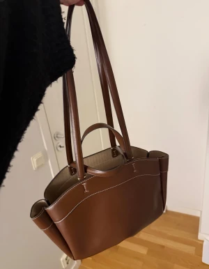 Brun shoppingväska i skinn - Stilren brun shoppingväska med vita sömmar och dubbla handtag. Väskan har en rymlig form och är tillverkad i slätt skinn, perfekt för att bära med sig allt man behöver. Snygg och enkel design som passar till många olika outfits.