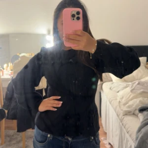 Svart stickad tröja med volang från JDY - Supersnygg svart stickad tröja från JDY med volang nedtill och puffade ärmar. Tröjan har ribbad struktur och rund halsringning, vilket ger en cool och trendig look. Perfekt till jeans eller kjol för en stilren outfit.