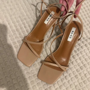 Beige sandaler med klack från NA-KD - Snygga beige sandaler från NA-KD med smala remmar och fyrkantig tå. Klacken är låg och stabil, perfekt för dig som gillar stilrena och minimalistiska skor. Remmarna går runt foten och fästs med spänne. Materialet är skinnimitation. Aldrig använda!