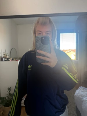 Blå Adidas vindjacka med gröna ränder - Säljer en blå Adidas vindjacka med ikoniska gröna ränder längs ärmarna och Adidas-logga på bröstet. Jackan har dragkedja framtill och sportig look, perfekt för dig som gillar streetstyle. Snygg passform och klassisk design. Står M men sitter som en L