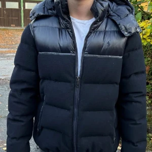 Marinblå pufferjacka från Polo Ralph Lauren - Säljer en marinblå pufferjacka från Polo Ralph Lauren med snygga glansiga detaljer upptill och på huvan. Jackan har dragkedja framtill, två sidofickor med dragkedja och en avtagbar huva. Perfekt för kalla dagar och har en riktigt clean look.