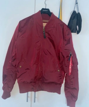 Vinröd bomberjacka Alpha Industries - Säljer en vinröd bomberjacka från Alpha Industries i storlek S. Jackan har klassisk 'Remove Before Flight'-tag på ärmen, ribbade muddar och dragkedja framtill. Tillverkad i slitstark polyester med orange foder och flera fickor. Perfekt för dig som gillar streetwear.
