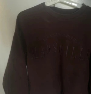 Mörkbrun sweatshirt med Marseille-tryck - Säljer en mörkbrun sweatshirt med upphöjt tryck där det står 'Marseille' på bröstet. Tröjan har rund hals och långa ärmar, perfekt för dig som gillar enkel och stilren streetwear. Materialet känns mjukt och skönt mot huden.