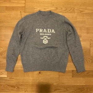 Grå stickad tröja från Prada - Snygg grå stickad tröja från Prada med vit logotyp och emblem på bröstet. Tröjan har rund hals, ribbade muddar och långärmad passform. Perfekt för dig som gillar exklusiva märken och vill ha en klassisk look med en modern touch.