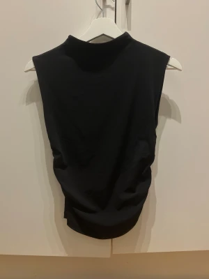 Svart ärmlös topp med rynk - Svart, ärmlös topp med hög hals och snygg rynkdetalj på sidan som ger en cool och modern siluett. Toppen är gjord i mjukt material och har en slim passform som framhäver figuren. Perfekt för dig som gillar minimalistisk stil med en twist.