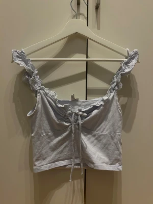 Ljusblå linnetopp med spets och snörning - Supersöt ljusblå linnetopp med volangprydda axelband i spets och snygg snörning framtill. Toppen är ärmlös och har en croppad passform som ger en fräsch vibe. Perfekt för varma dagar och chill häng med vänner.