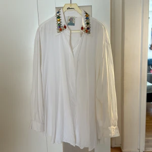 Vit skjorta med pärlor från Mama - Vit oversized skjorta från Mama med långa ärmar och en krage dekorerad med stora färgglada pärlor och stenar. Skjortan har en luftig passform och är tillverkad i ett mjukt, lätt material som ger ett snyggt fall.