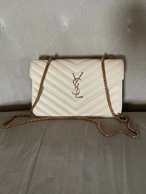 Beige quiltad YSL axelväska med kedja - Snygg beige axelväska från YSL med quiltad design och guldfärgad kedja. Framsidan har YSL-logga i metall och baksidan ett insytt hjärta. Väskan är tillverkad i skinn och har dragkedja på insidan. Perfekt för dig som gillar stilrena detaljer och exklusiv känsla.