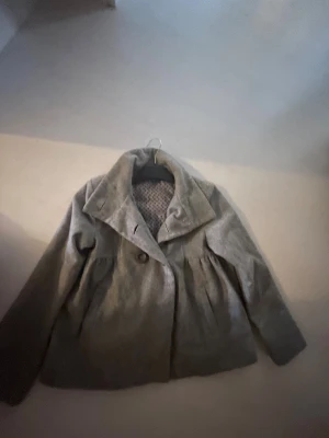 Grå kappa med dubbelknäppning - Snygg grå kappa med bred krage och dubbelknäppning framtill. Jackan har ett lätt utställd passform och långa ärmar. Insidan har ett diskret mönstrat foder. Perfekt för dig som gillar stilrena och klassiska vibes.