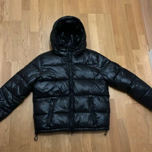 Svart pufferjacka från Lexington - Snygg svart pufferjacka från Lexington med huva och dragkedja framtill. Jackan har två fickor med dragkedja och är en modern stils jacka. Nypris:2500 kr 