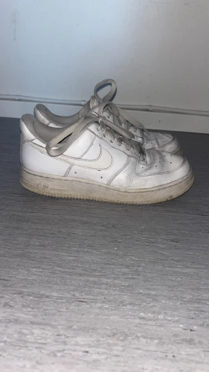 Nike Air Force 1 vita sneakers - Klassiska Nike Air Force 1 sneakers i helvitt skinn med rund tå och snörning. Modellen har den ikoniska swoosh-loggan på sidan och en platt sula. Perfekta för dig som gillar streetstyle och tidlös design. Några fläckar som inte går bort.