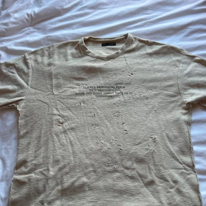 Beige t-shirt med text från Zara - Beige t-shirt från Zara i storlek S med korta ärmar och tryckt text framtill. T-shirten har en rå och sliten look med små hål och fransiga kanter, vilket ger en edgy vibe. Materialet är mjukt och känns som bomull. Perfekt för dig som gillar streetstyle.