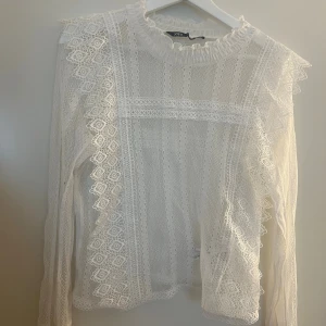Vit spetsblus med volang från Zara - Säljer en vit blus från Zara med transparent spets och dekorativa volanger vid axlar och halsringning. Blusen har långärmade ärmar och ett romantiskt mönster med broderade detaljer. Perfekt för dig som gillar en feminin och trendig look.