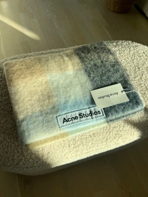 Rutig halsduk Acne Studios i ull - Snygg och fluffig halsduk från Acne Studios med rutigt mönster i ljusblått, beige och grått. Tillverkad i Italien av mjuk ull, perfekt för att ge din outfit en cool touch. Acne Studios-logga är tydligt synlig på etiketten.