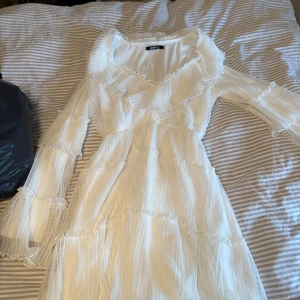 Vit volangklänning från Gina Tricot - Supersöt vit klänning från Gina Tricot med volanger och transparenta detaljer. Klänningen har långärmade ärmar och är gjord i ett lätt, plisserat tyg. Perfekt till t.ex skolavslutning