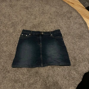 Mörkblå jeanskjol från Brooker - Snygg mörkblå jeanskjol från Brooker med klassisk femficksdesign, dubbla knappar och bälteshällor. Kjolen är i kort modell och har bakfickor med dekorativa sömmar. Storlek 170