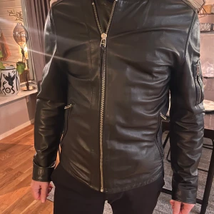 Svart skinnjacka Tiger of Sweden - Snygg svart skinnjacka från Tiger of Sweden Jeans i storlek Large. Jackan har raka linjer, silverfärgade dragkedjor fram och på fickorna samt en extra ficka på ärmen. Klassisk och stilren modell med lång ärm och normal passform. Fint tunnt svart skinn. Använd ett fåtal gånger. Nypris 6000kr