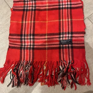 Rutig röd halsduk från Burberry - Säljer en klassisk rutig halsduk från Burberrys i rött med inslag av svart, vitt och gult. Halsduken är tillverkad i 100% lammull och har snygga fransar i ändarna. Perfekt accessoar för att lyfta din outfit med lite brittisk vibe.